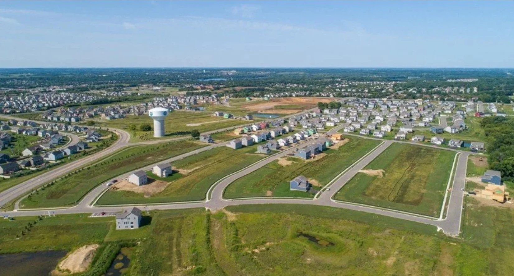 Aerial view of a new residential neighborhood with houses under construction, green open fields, and roads, with a water tower in the background.