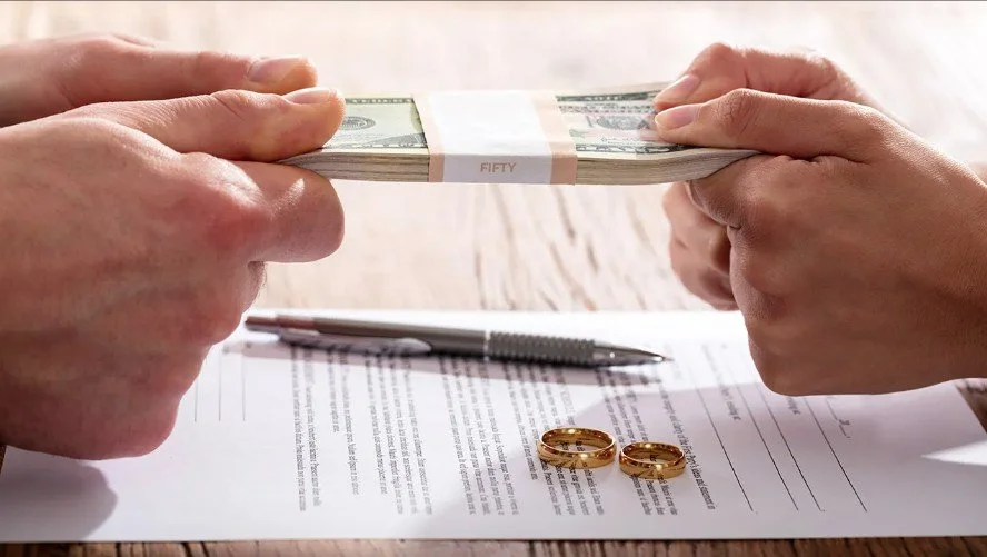 Two hands exchanging a stack of fifty dollar bills over a document with two gold wedding rings, a pen, and some text on a wooden surface.