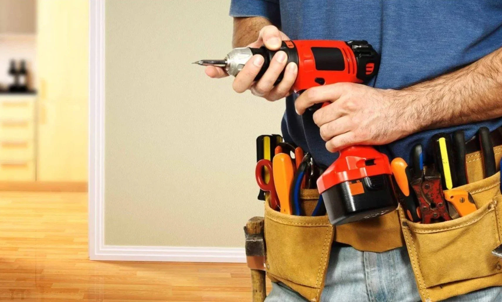 A person wearing a tool belt with various tools, holding a cordless screwdriver, standing in an indoor setting with wooden flooring and a beige wall.