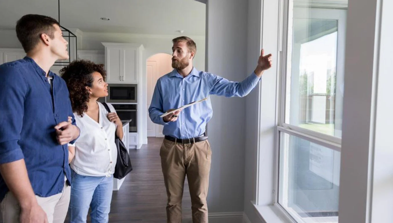 Real estate agent showing a house to a couple, pointing at the window inside a modern kitchen.