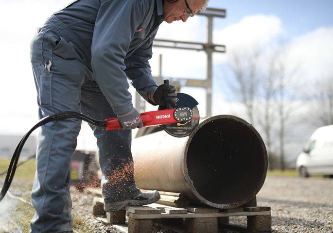 Man wearing gray work coveralls using a handheld metal cutting tool on a large pipe outdoors, sparks flying as he cuts the pipe.