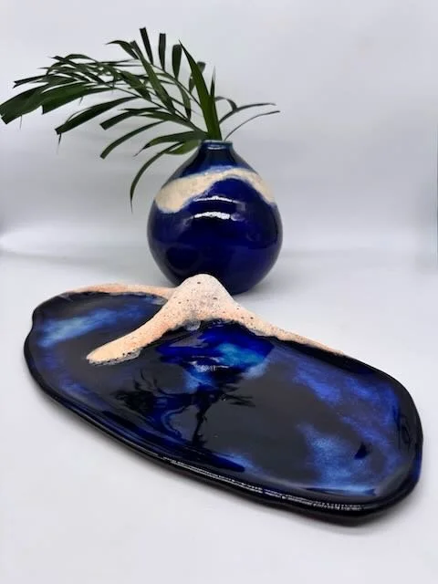 A ceramic vase with a green palm leaf, and a black and blue decorative tray resembling Mount Fuji with white volcanic ash.
