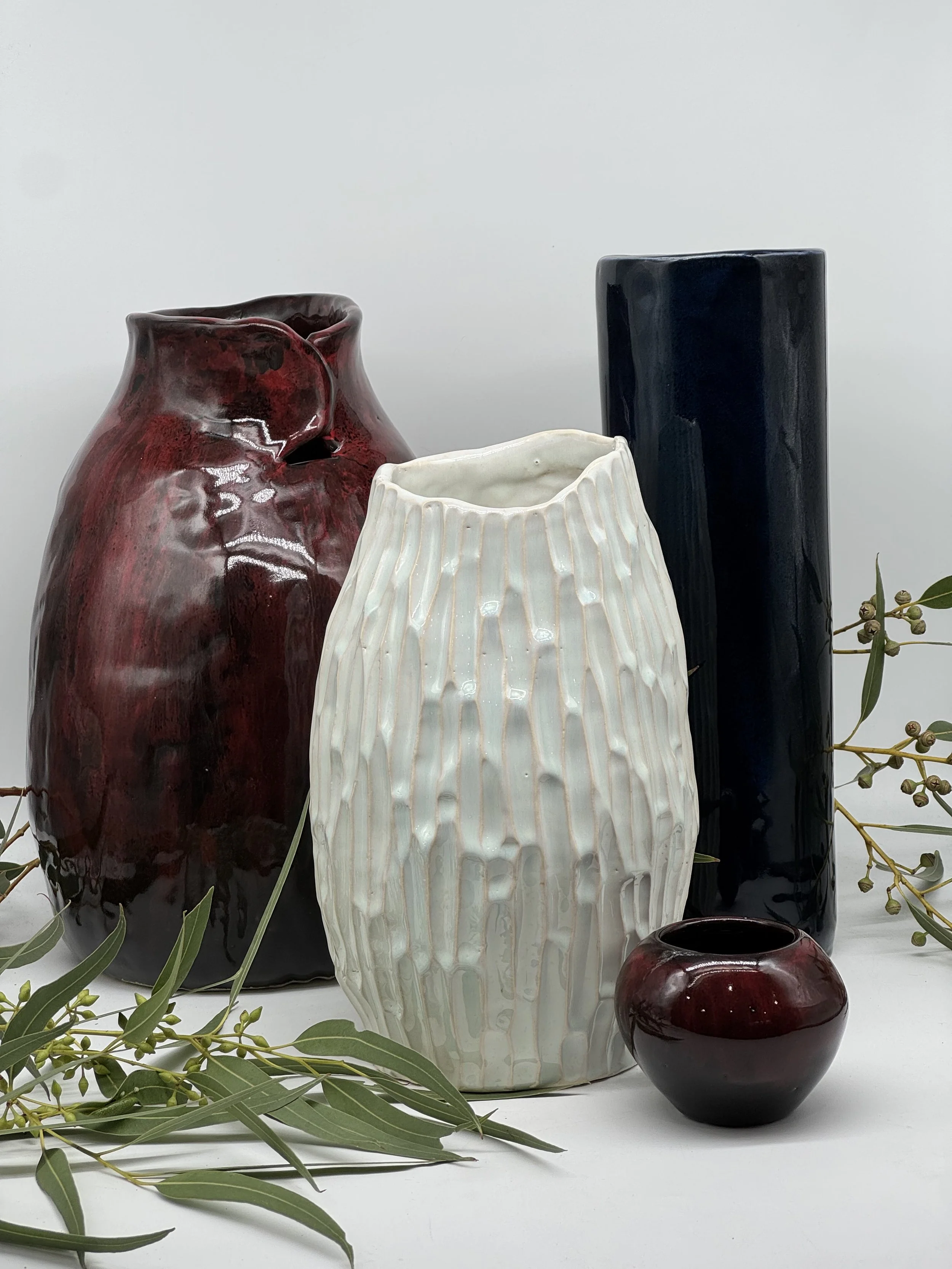 Arrangement of five ceramic vases in various shapes, sizes, and colors, with greenery and small branches in the foreground, on a white surface against a light background.