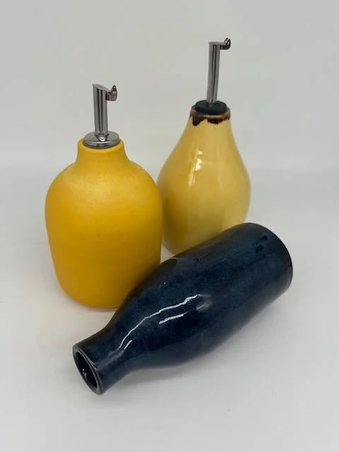 Three ceramic oil dispensers with metal pour spouts, two are yellow and one is dark blue, placed on a white surface.