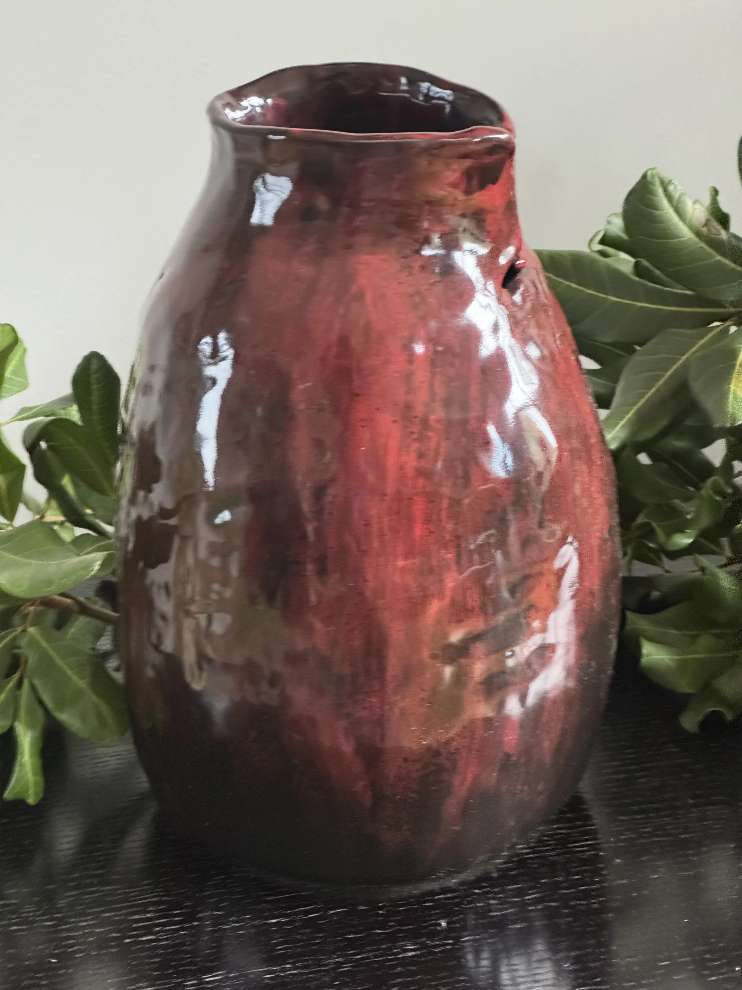 A glazed red and black ceramic vase on a black surface with green leaves in the background.