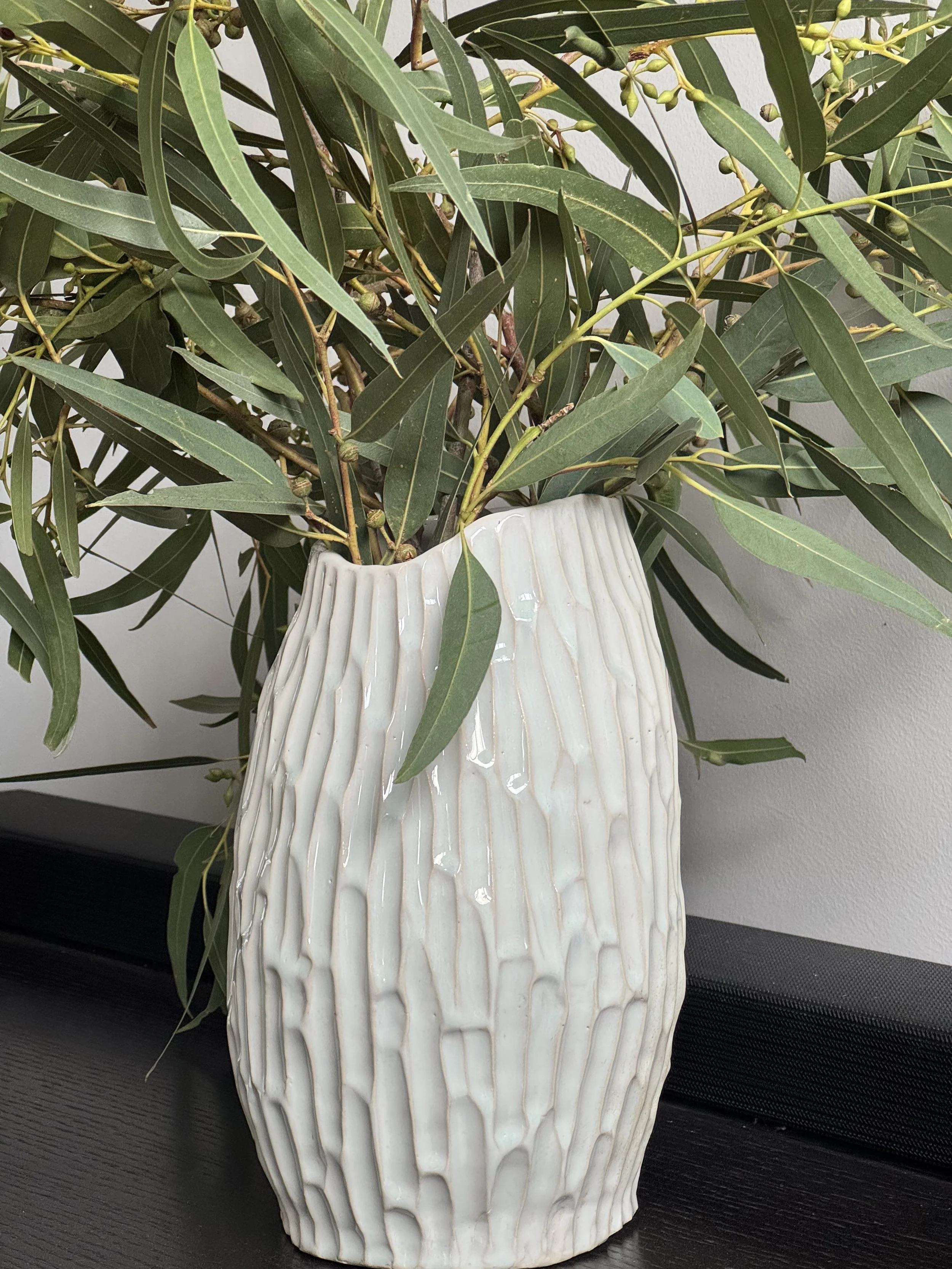 A white ceramic vase with vertical ridges, filled with green leafy branches with small green buds, placed on a black surface against a white wall.