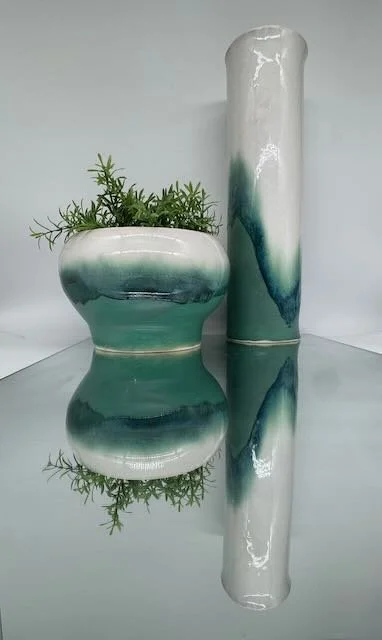Ceramic vase with green plant and a tall, elongated ceramic vase with green and white glaze, reflected on a mirror surface.