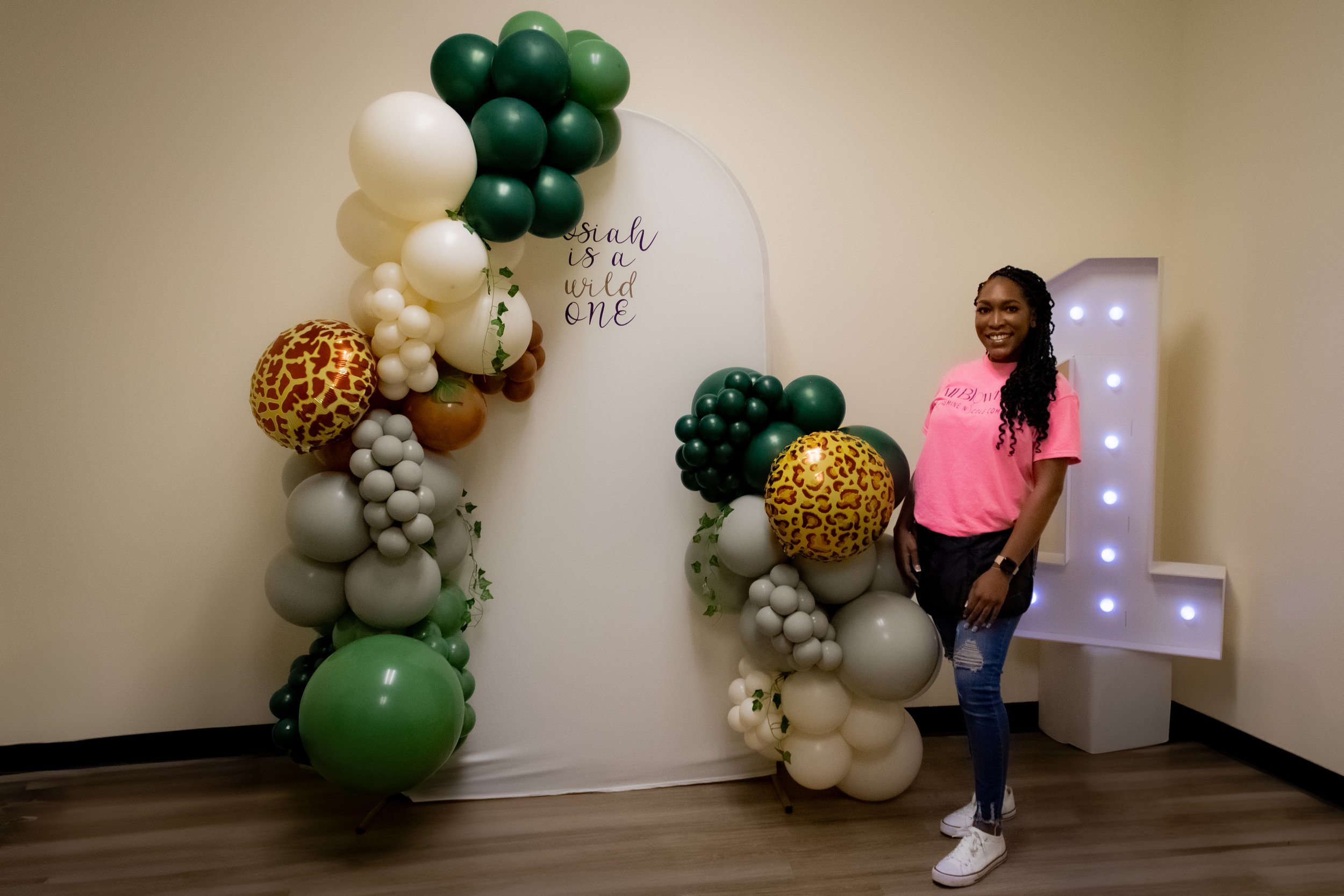 Woman standing beside a balloon arch with animal print and jungle colors, with a backdrop saying "is a wild one" and a large illuminated number one.