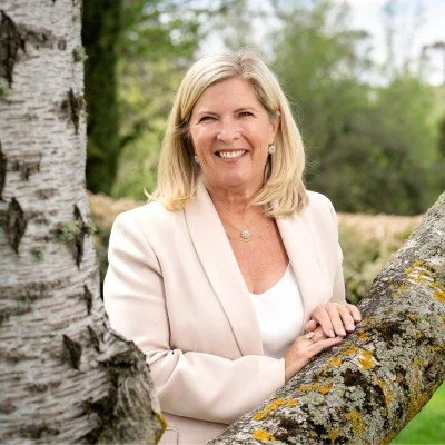 A smiling woman with blond hair wearing a light-colored blazer, standing outdoors among trees.
