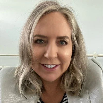 A woman with shoulder-length, wavy blonde hair smiling at the camera, wearing a light-colored blazer and a striped top, sitting indoors with a white wall and a lightning cable behind her.