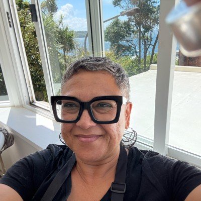 Person with short gray hair wearing large black glasses smiling indoors near a window with a view of trees and water.