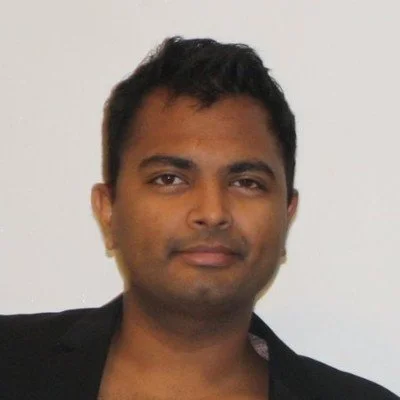 A man with short black hair and brown skin, wearing a black blazer, looking at the camera against a plain light-colored background.