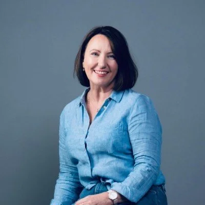 A woman with shoulder-length dark hair smiling and wearing a light blue button-up shirt against a gray background.