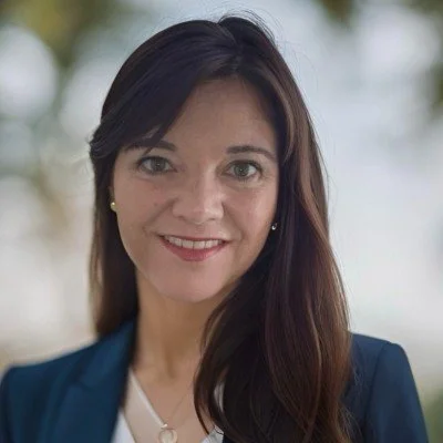 A woman with long dark hair wearing a navy blazer and white top, smiling outdoors with blurred trees in the background.