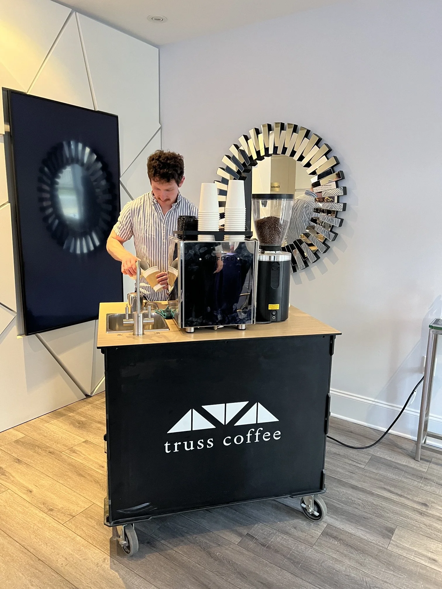 A mobile coffee cart labeled 'truss coffee' with a person pouring coffee and a coffee grinder on the counter.