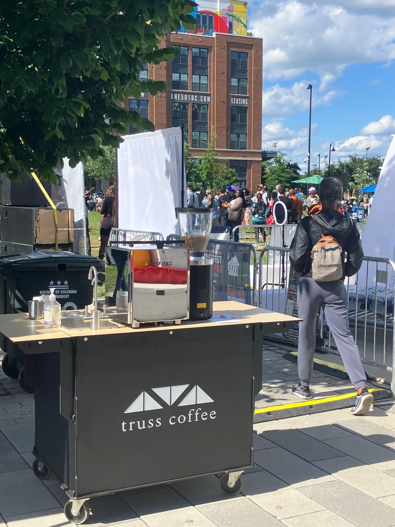 A busy outdoor scene with a coffee cart labeled 'truss coffee' in the foreground. A person in gray sweatpants, a black jacket, and a backpack is walking away from the camera. There are many people in the background gathered in what appears to be a park or event space, some with umbrellas and hats. A brick building with windows and signs, including one with a website URL and the words 'LEASE' and 'ONE 501DC.COM,' is visible under a partly cloudy sky.