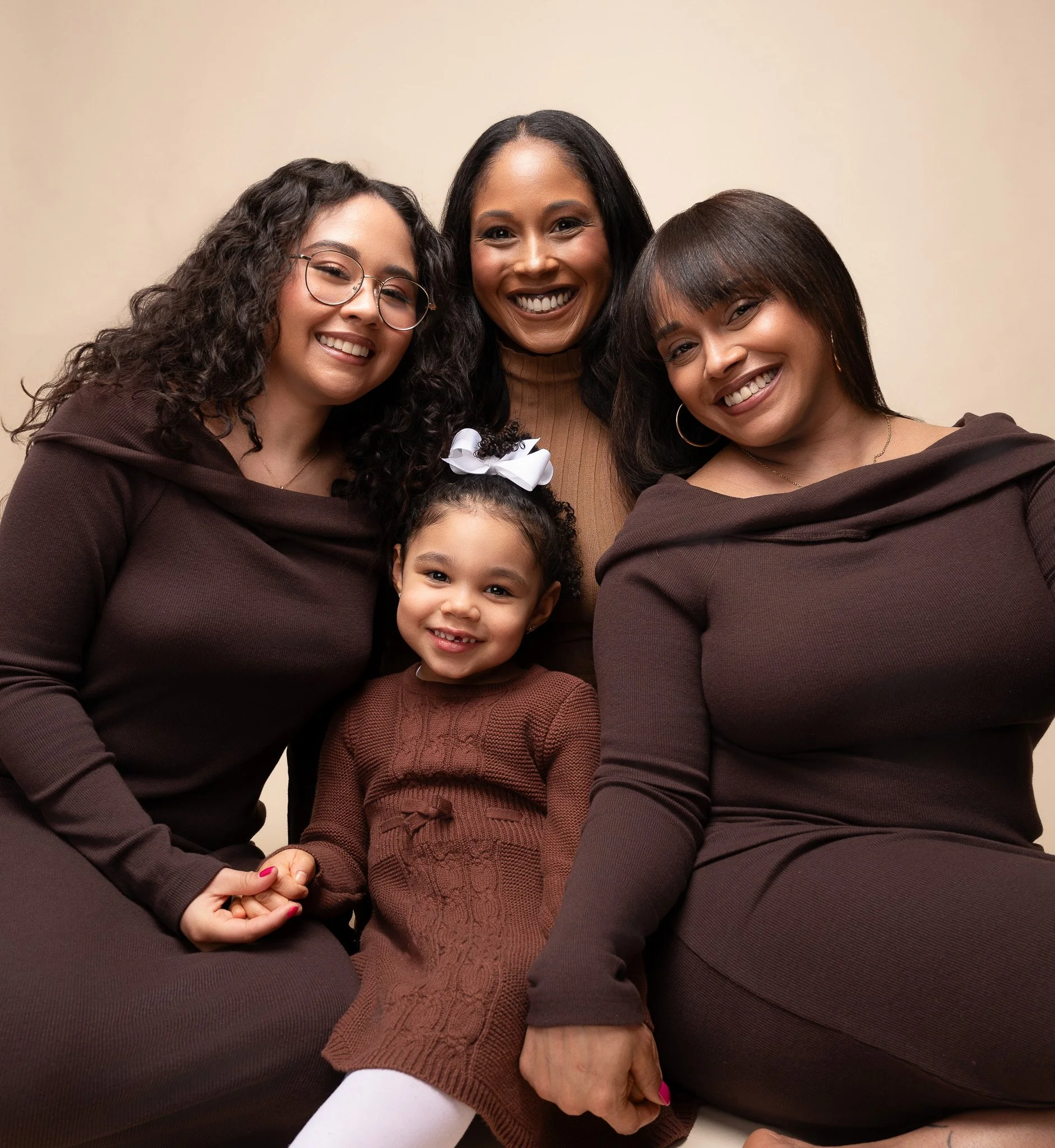 A group of four women and one young girl smiling and sitting close together against a plain background.