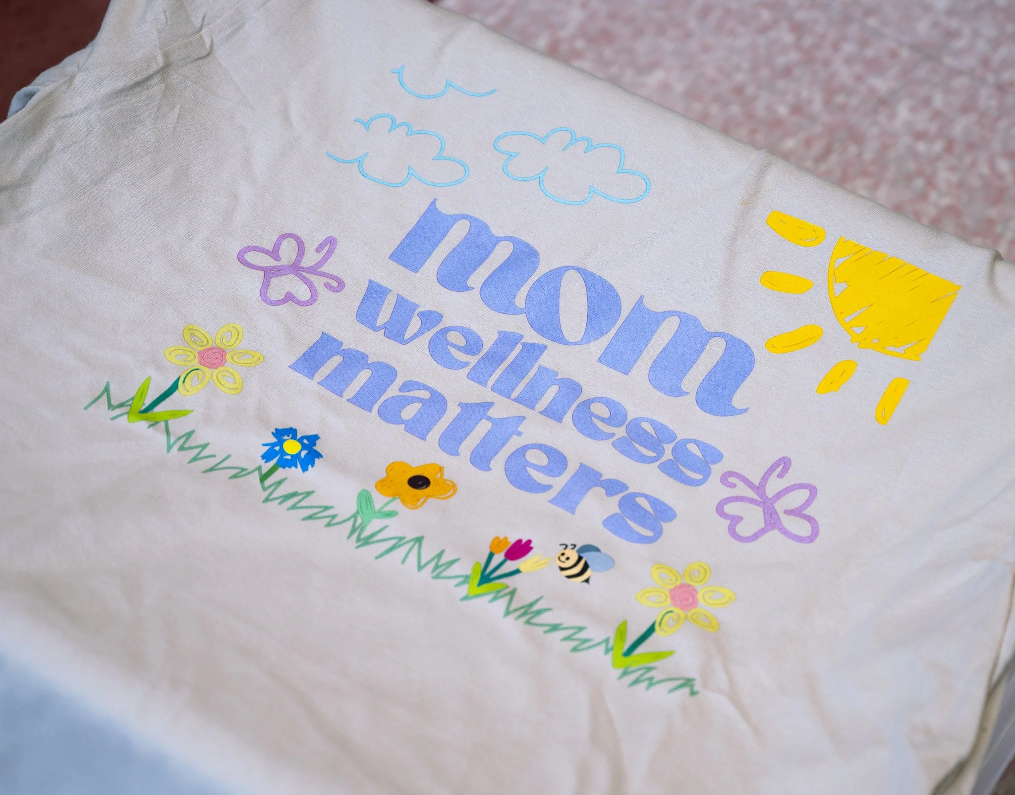 A white surface with a colorful drawing of the sun, clouds, flowers, a bee, and the words "Mom Wellness Matter" in purple and blue letters.
