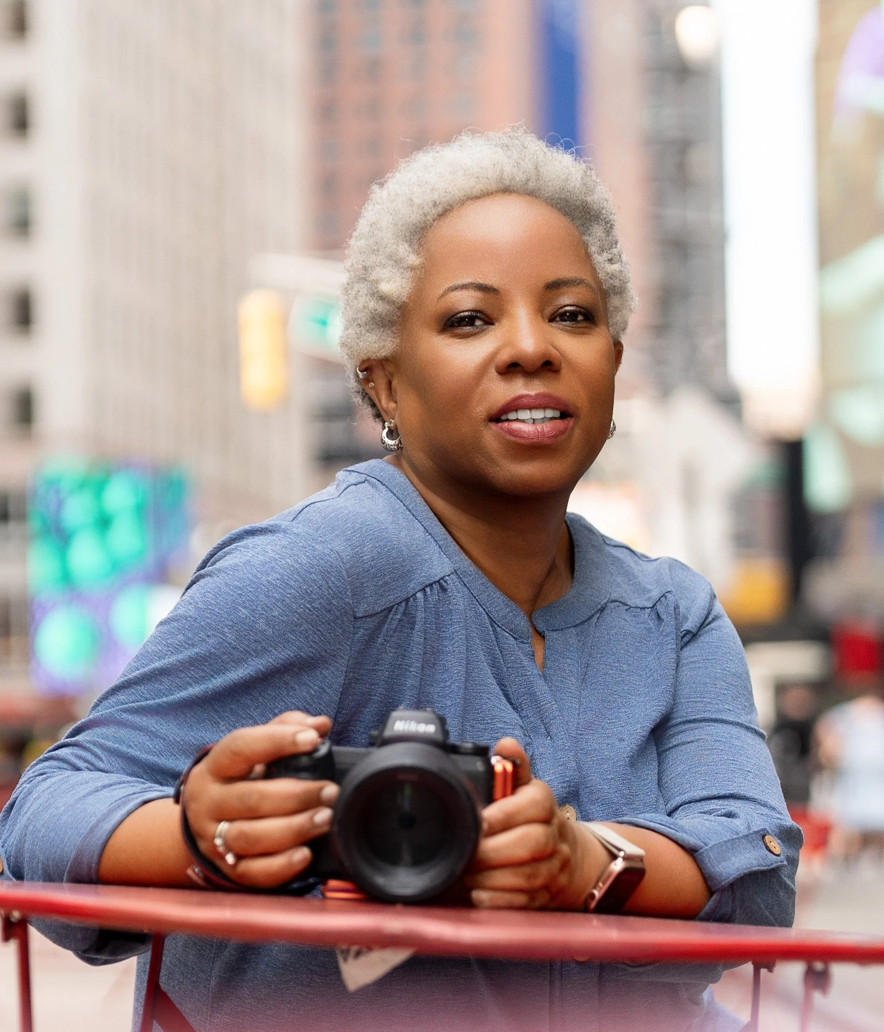 A woman with short gray hair holding a camera on a city street with tall buildings and blurred pedestrians in the background.