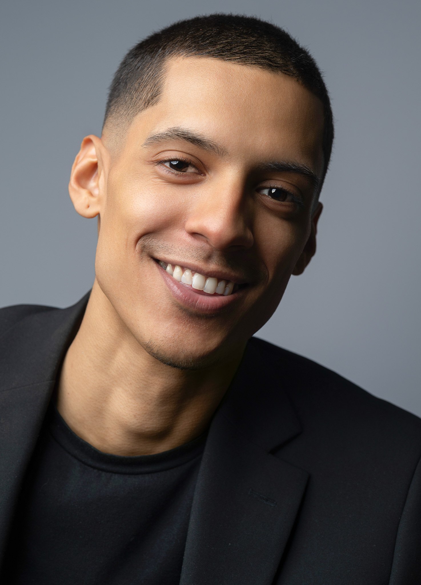 Close-up portrait of a smiling young man with short dark hair, wearing a black blazer and black shirt, against a gray background.