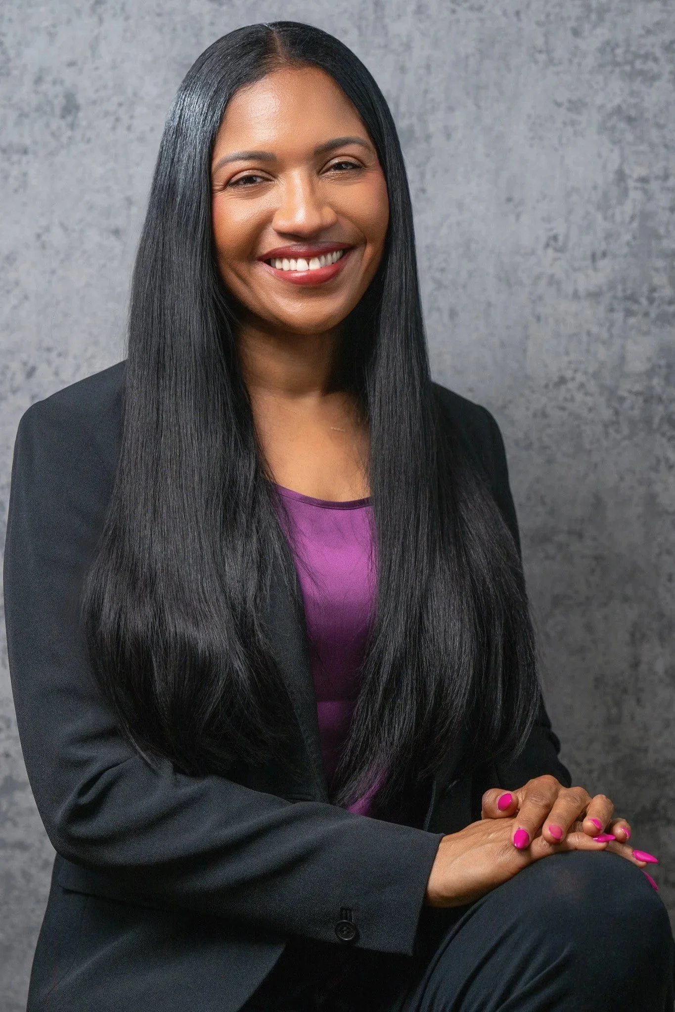 A smiling woman with long black hair, wearing a purple top and a black blazer, sitting against a gray textured background.