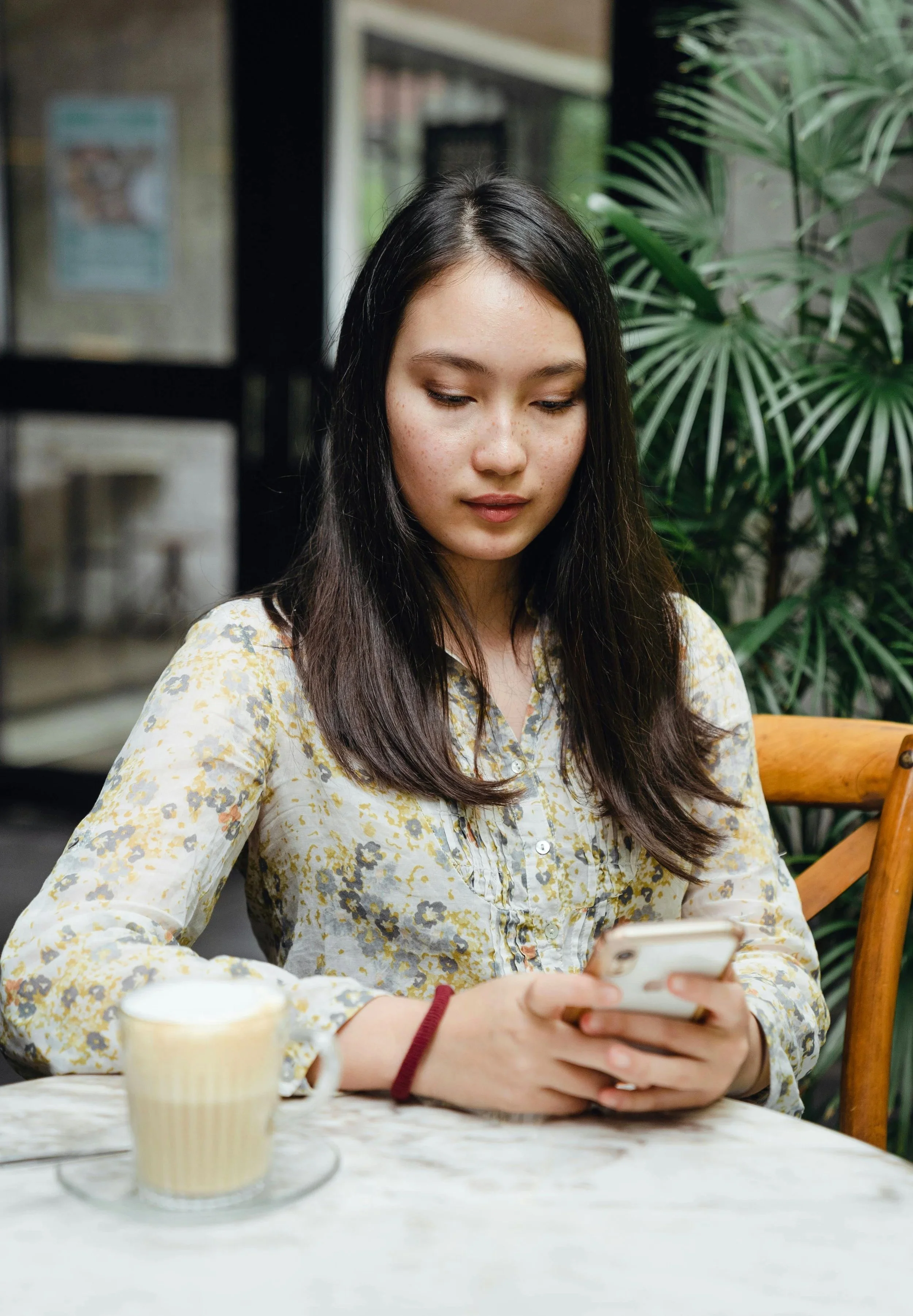 A young woman with long dark hair and a floral shirt sitting at a table in a cafe, looking at her phone with a coffee mug nearby, greenery in the background.