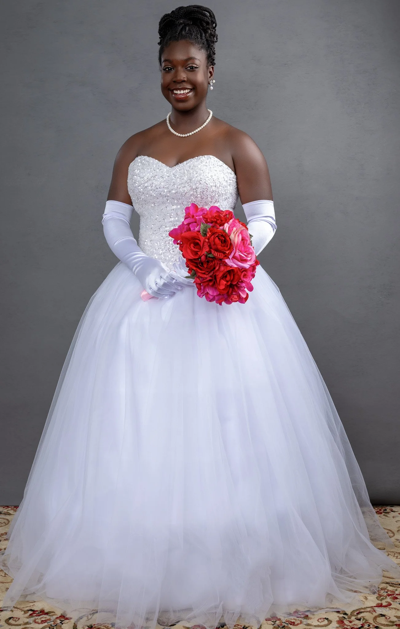 A young woman in an elaborate white dress holding a bouquet of pink and red roses, smiling, against a gray background.