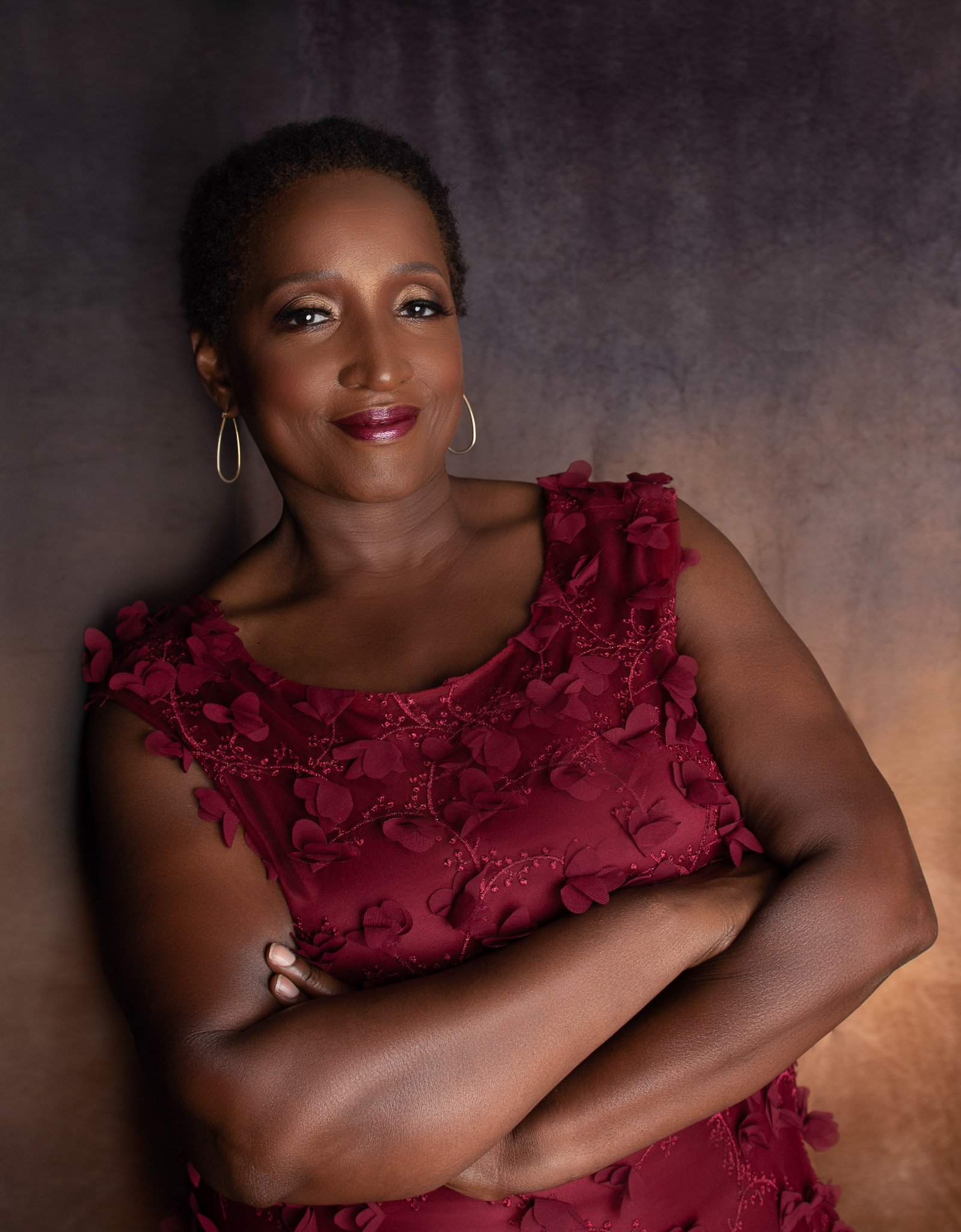 Portrait of a woman with short curly hair, wearing gold hoop earrings and a burgundy dress with floral embroidery, standing against a gradient background.