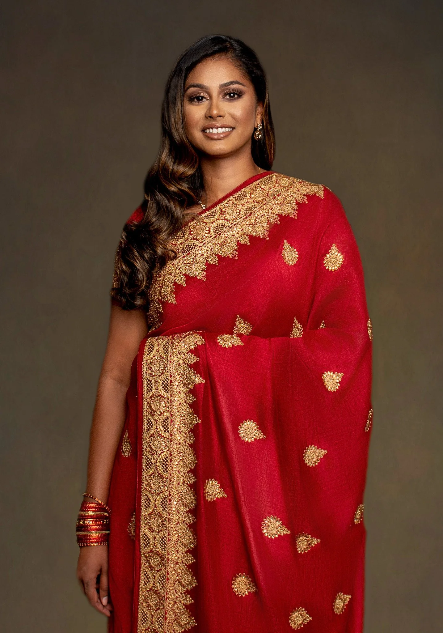 A woman wearing a red saree with gold embroidery and details, standing against a neutral background.
