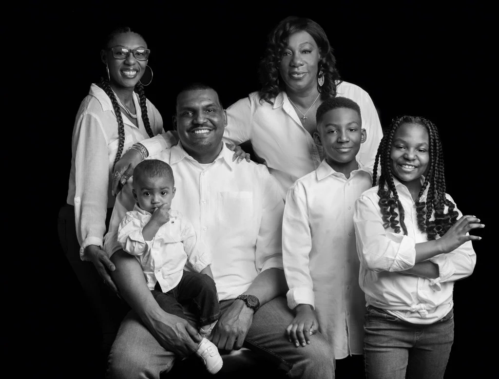 Black and white studio portrait of a family of six, with parents and four children smiling together while wearing coordinated white shirts against a black background