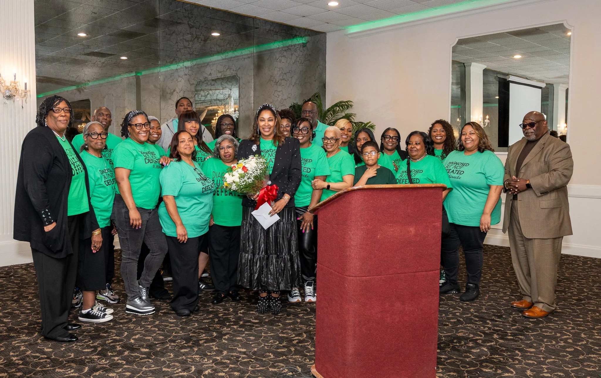 A group of people standing together indoors, some wearing matching green t-shirts, with one woman holding a bouquet of flowers, posing for a photo.
