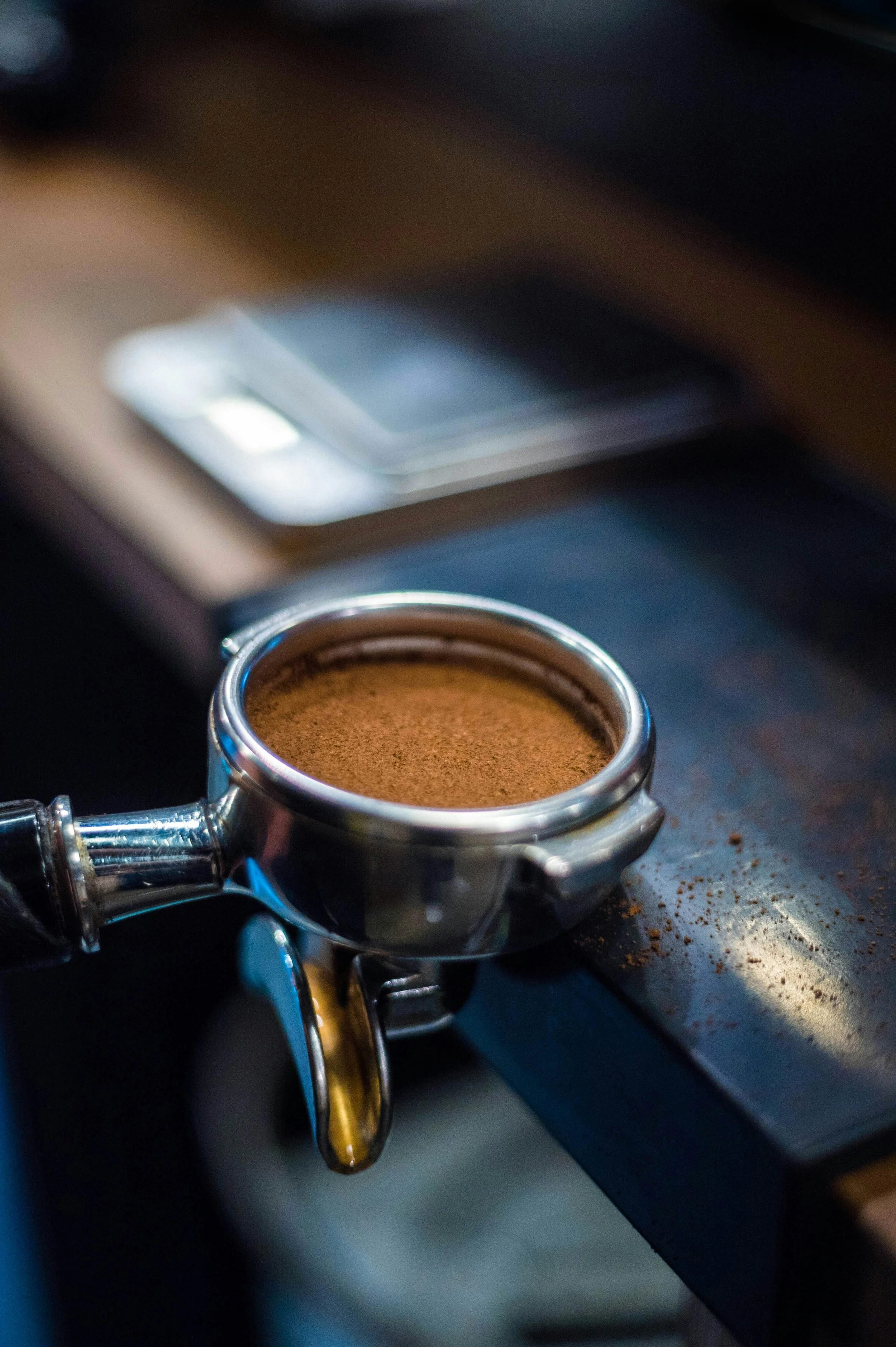 Close-up of an espresso machine portafilter filled with ground coffee on a dark countertop with spilled coffee powder.