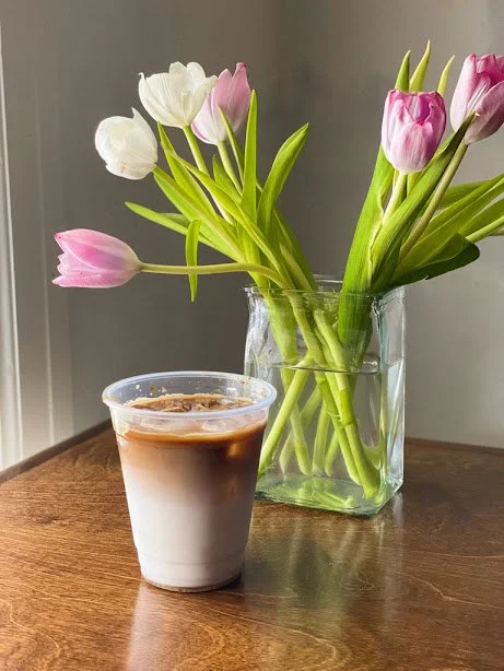 A glass vase filled with pink and white tulips on a wooden surface, with a plastic cup of iced coffee in front of it.
