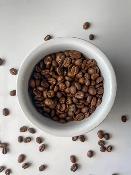 Coffee beans in a white bowl with some beans scattered around on a light gray surface.