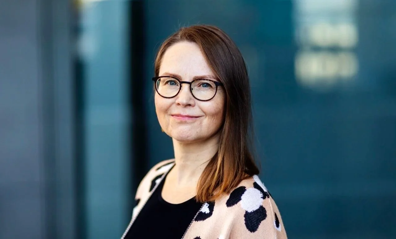 Noora Jokinen hymyilee katse kameraan yllään tumma paita ja persikan värinen, pantterikuvioinen neuletakki.