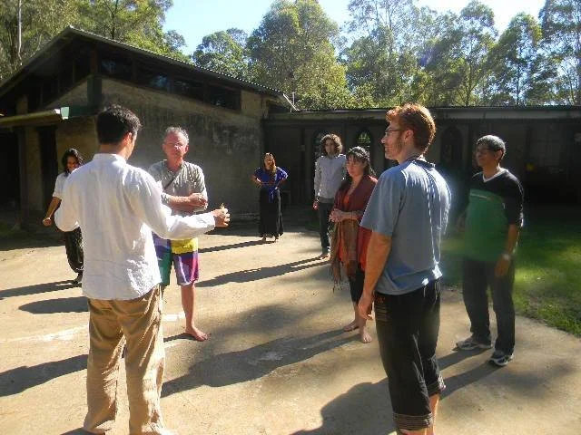 Group of people standing outdoors, with a man in a white shirt and tan pants speaking to the group. Some individuals are dressed casually, and there is greenery and a building in the background.
