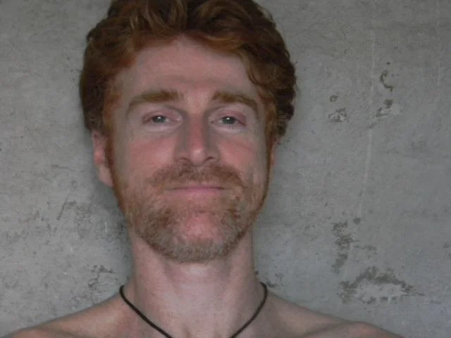 A man with red hair and beard smiling at the camera against a gray concrete wall.