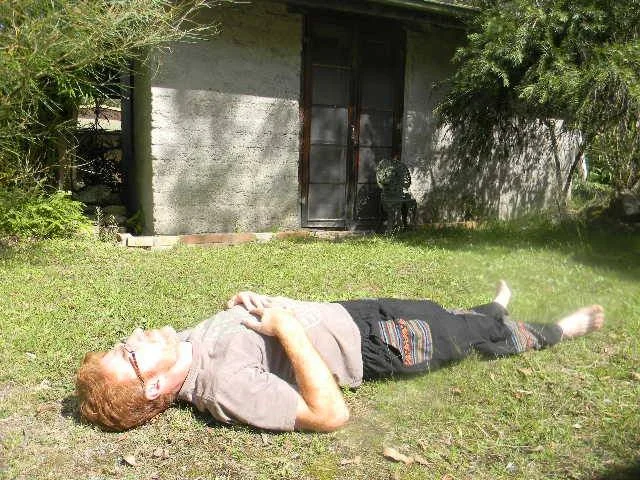 An elderly man lying on his back on a grassy yard, relaxing and enjoying the sunshine, with a small building and trees in the background.
