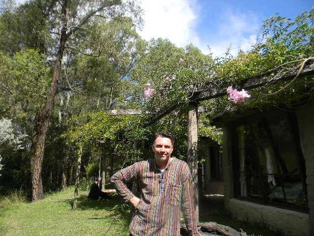 A smiling man standing outdoors in a garden with trees and pink flowers, next to a house with large windows and a pergola with pink flowering vines.