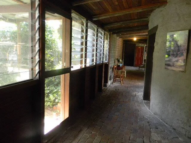 Interior of a rustic house with a brick floor, wooden ceiling, and large glass windows with louvered shutters, featuring a small table and chairs.