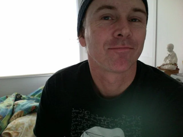 A man taking a selfie indoors, wearing a dark T-shirt and a backwards cap, smiling slightly, with a bed and a decorative doll in the background.