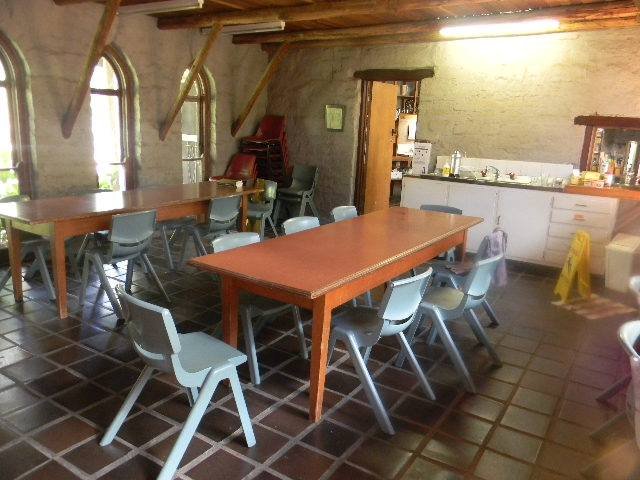 A room with a tiled floor, wooden beams, and large arched windows. There are several picnic-style tables with blue chairs around them. In the background, there is a kitchenette with white cabinets and a counter with various items, and a wet floor sig