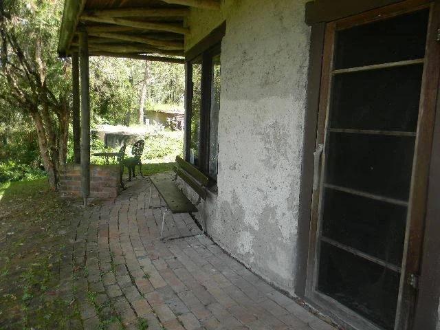 Porch with brick floor, peeling white wall, metal bench, black chair, and large window, overlooking greenery and trees outside.