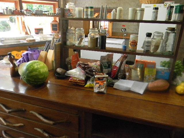 Kitchen countertop with a large watermelon, various spice jars, seasoning packets, a wooden cutting board, and some fruit, with kitchen shelves holding bottles and containers in the background.