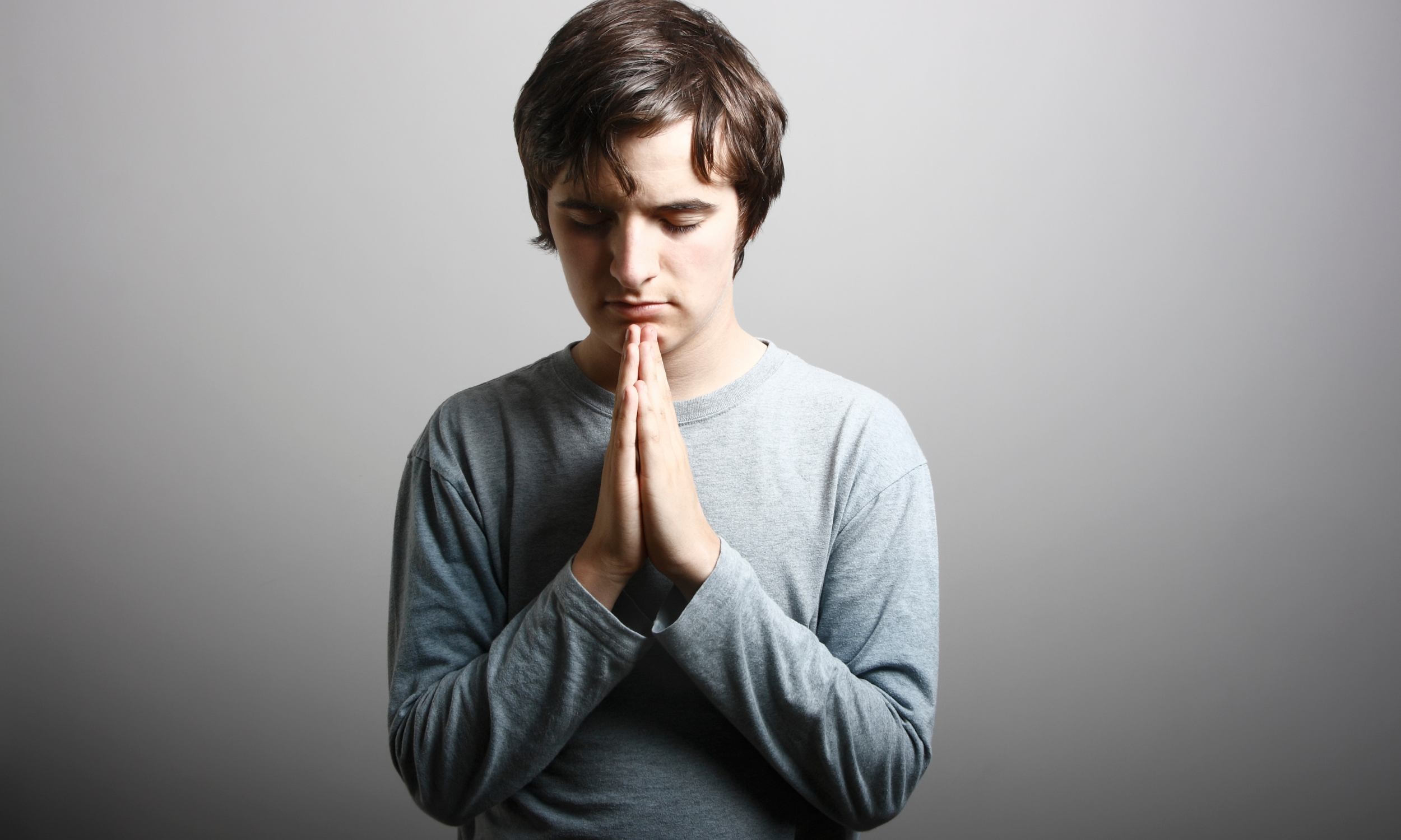 A young man with dark hair, wearing a gray long-sleeve shirt, has his eyes closed and hands pressed together in a prayer or meditative pose against a plain gray background.