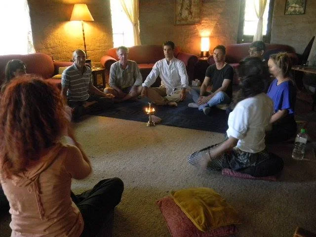 Group of seven people sitting cross-legged on the floor in a circle, in a cozy room with warm lighting, participating in a meditation or spiritual practice session.