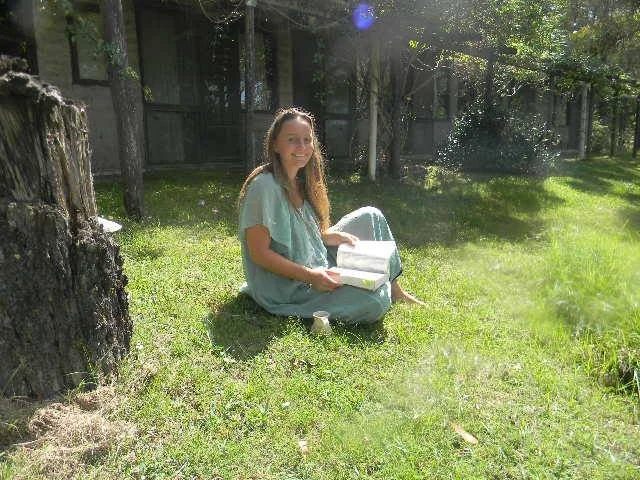 A woman with long hair, dressed in a greenish-blue top, sitting cross-legged on grass outdoors, smiling, with a box in her hands. There is a tree trunk on the left, and a fence with trees in the background.