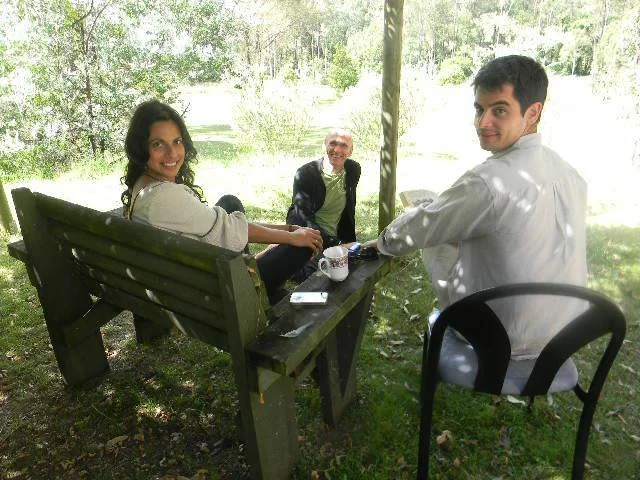 Three people sitting outdoors around a table with mugs, with trees and grass in the background.