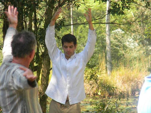 Three men stand outdoors in a forest, with one man in a white shirt in the center raising his hands, two other men in striped shirts gesturing towards him.