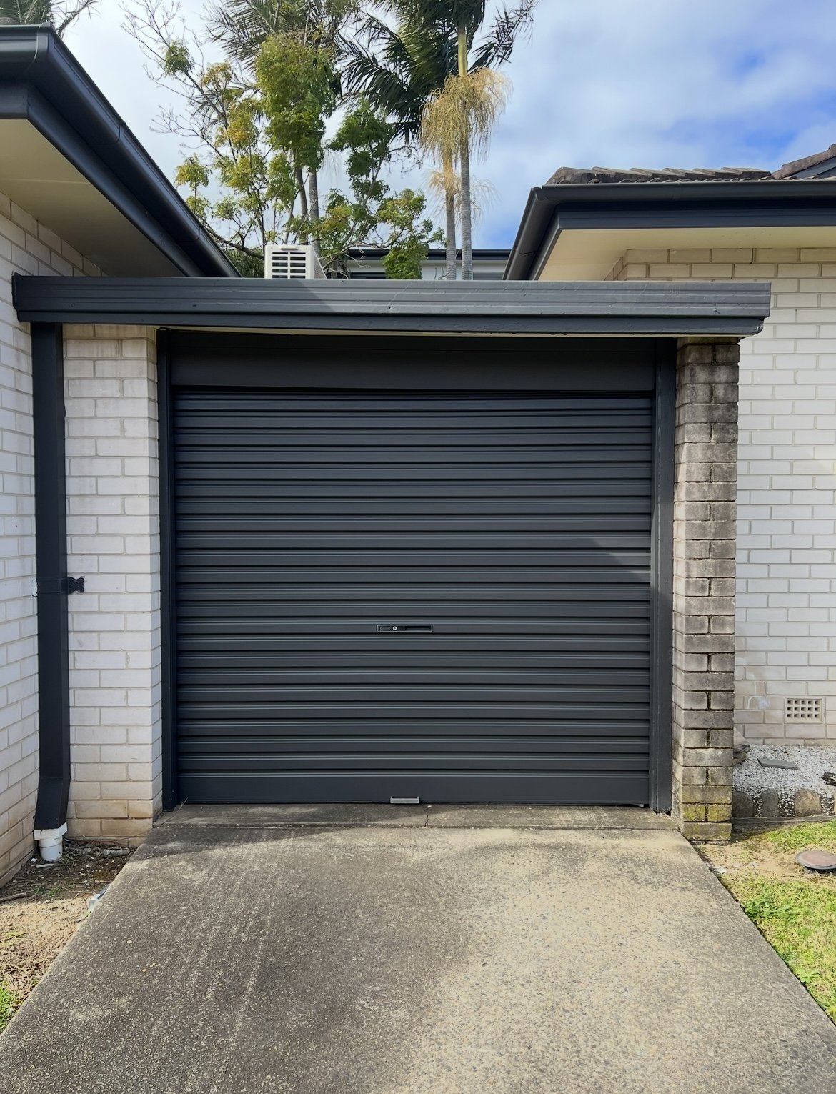 Closed black metal roll-up garage door after our repaint at a strata property. On a house with brick and cement walls, a concrete driveway, and trees in the background.